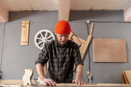 A young man in a orange hat  carpenter builder in work clothes processing a wooden board with a...