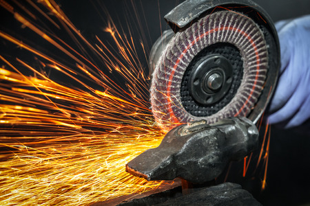 Close-up on the sides fly bright sparks from the angle grinder machine. A young male welder in a white working gloves grinds a metal product with angle grinder in the garageの写真素材