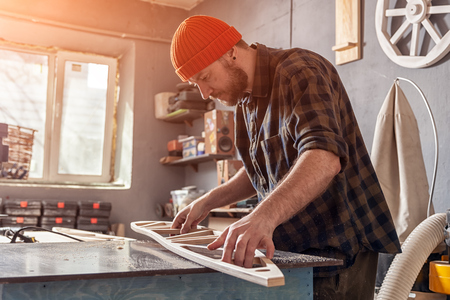 A young man carpenter builder in work clothes processing a wooden board with a milling machine in the workshop, around a lot of equipment, wooden boards. Home repair concepts.の写真素材