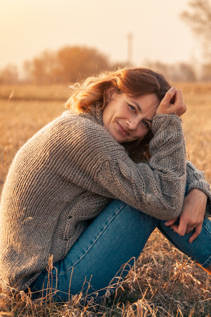 Portrait of a beautiful young model in warm clothes enjoy day, on background field in  sunny autumn day . Autumn warm photo. Woman smiling and look away, joyful cheerful mood.の写真素材