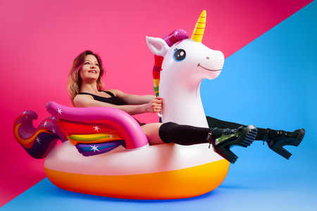 Concept summer mood, relaxation and beauty.Modern portrait of a young woman in a yellow swimsuit, bright socks and sandals resting on an inflatable unicorn mattress on an isolated pink-blue backgroundの写真素材