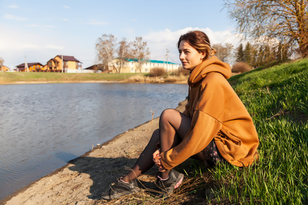 A beautiful woman in a warm sweatshirt, dress and sneakers is sitting on the shore of the lake, resting, enjoying nature, looking at the water on a warm summer dayの写真素材