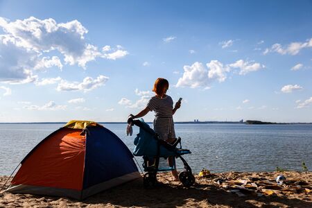 A young mother rolls her newborn baby in a stroller and looks into the phone on a warm summer day on the sea with a sandy beach. Family vacation at seaの写真素材