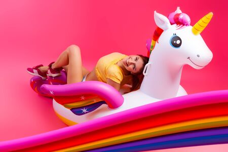 Summer fashionable girl, pink and joyful mood. Beautiful slim woman in a summer clothes, having fun and enjoying life on a white inflatable unicorn on a pink isolated backgroundの写真素材
