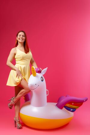 Summer Fashion  beautiful woman in summer clothes having fun, smilling and posing  with balloons On Unicorn Float on isolated pink backgroundの写真素材