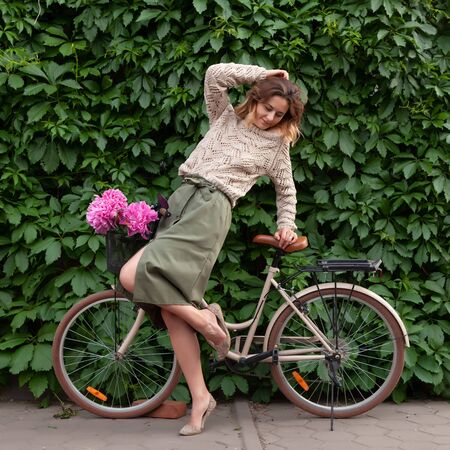 A young dark-haired woman in sweater and a beige skirt  sitting on beige retro bicycle  with bouquet of pink peonies in a wicker basket on the background of a vine-covered wallの写真素材