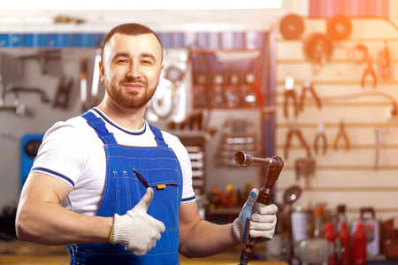 A young man-mechanic in overalls poses at his workplace and holds a pneumatic wrench in his hand, in...