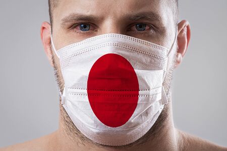 Young man with sore eyes in a medical mask painted in the colors of the national flag of Japan. Medical protection against airborne diseases, coronavirus. Man is afraid of getting the fluの写真素材