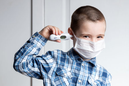 Young boy with sore eyes in a medical mask measures the temperature of a thermometer in his ear....