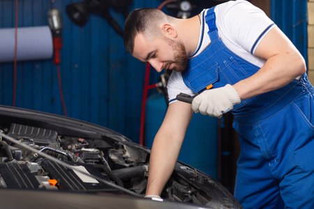Handsome young male auto mechanic in special uniform clothes holding a flashlight, looking for...