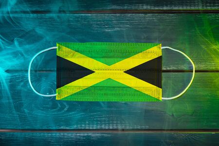 Medical mask for protection against airborne diseases, painted in the national flag of Jamaica  on a black background in blue-green smoke. Medical protection against airborne diseases, coronavirusの写真素材