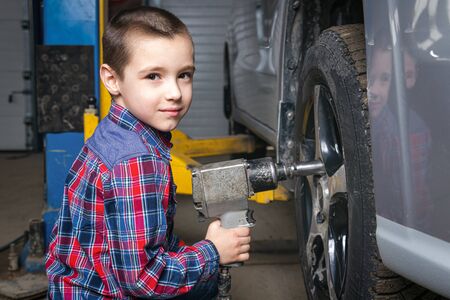 A young boy, a young auto worker, makes a tire change with a pneumatic wrench in the garage of a service station. A child learns the mechanics changing profession in auto repair service.の写真素材