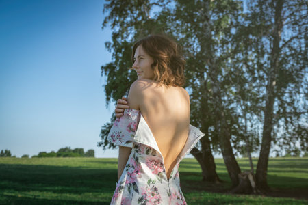 A beauty attractive woman in a light dress with bare shoulders and a floral print stands with her back to the camera with her dress zipped open. The concept of loneliness, thoughtfulness and longing for a woman. Close-up of a female bare backの写真素材