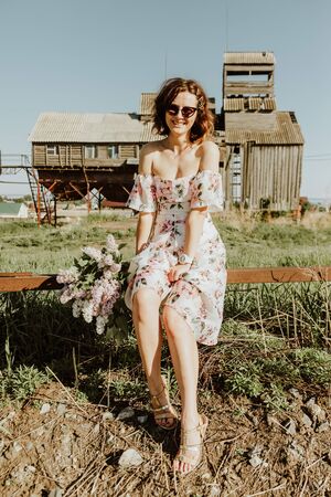 Beauty woman in a romantic light dress with bare shoulders and a hat  sitting and looking cheerfully at the camera, in the background an old abandoned village and green grass. The concept of life in the countryside in the villageの写真素材