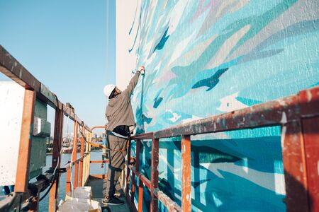 Male professional painter builder paints the wall of a new building at high altitude in a construction cradle against a background of cityscape and a riverの写真素材