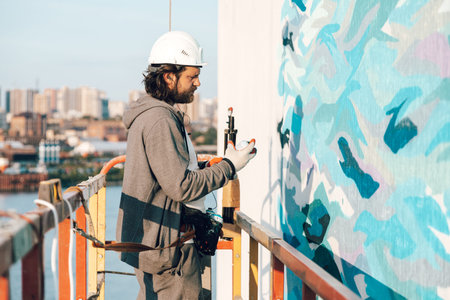 Contractor, artist at high altitude in a building cradle performs facade painting, decorating and...
