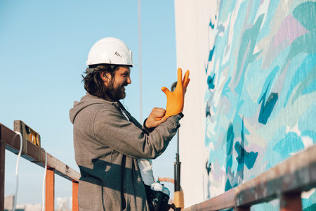 Contractor, artist at high altitude in a building cradle puts on gloves and prepares to carry out facade painting, decorating and home renovation against the backdrop of the city landscape and the riverの写真素材