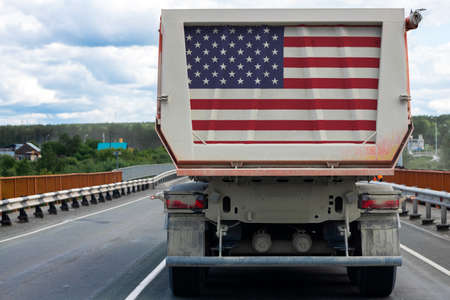 Big truck with the national flag of USA moving on the highway, against the background of the village and forest landscape. Concept of export-import, transportation, national delivery of goodsの写真素材