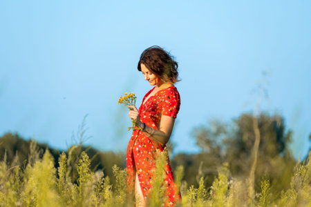 A dark-haired woman smiles, walks along the green field dressed in romantic dress on a summer day....