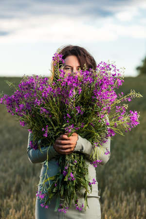 Portrait of a beautiful young woman in blue romantic dress with large bouquet of purple flowers walking on the field, enjoying the day, on a sunny summer day. The concept of the unity of the child and nature, peaceful mood, environmental lifeの写真素材