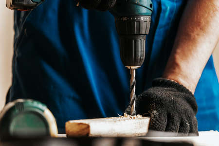 Close-up of a male construction worker in a black dirty construction overall closes a screw with a black modern screwdriver in a wooden block in the workshopの写真素材
