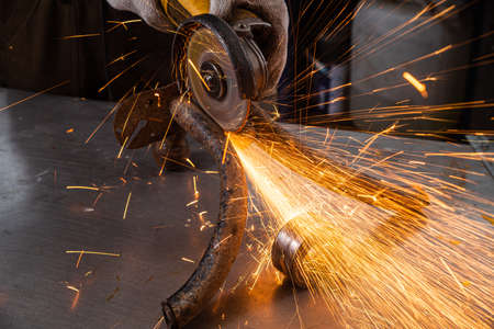 Close-up on the sides fly bright sparks from the angle grinder machine. A young male welder in a blue working gloves grinds a metal product with angle grinder in the garageの写真素材