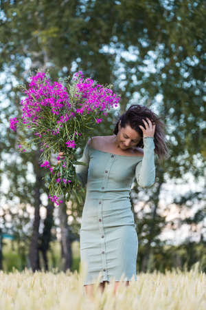 Portrait of a beautiful woman with a bouquet of flowers walking in the fields, in background field and forest. The concept of the unity of women and nature, peaceful mood, eco-friendly lifeの写真素材