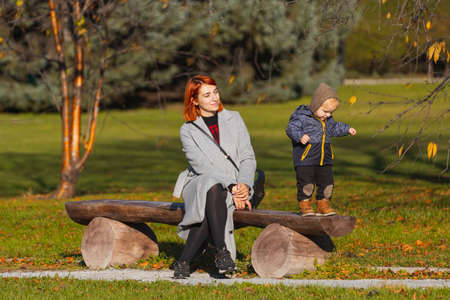Mom helps her little son take his first steps on a wooden park bench on a warm summer day. Happy childhood conceptの写真素材