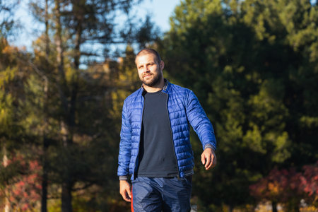 Young man in warm autumn clothes, blue coat, t-shirt enjoys the warm sun and walks in a city park on...