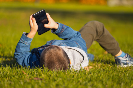 A cheerful boy in a denim jacket lies on a bench and plays with a smartphone in a city park. Concept...