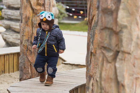Adorable little 1-2 year old toddler boy having fun on the playground, the child wears a blue jacket with an owl hatの写真素材