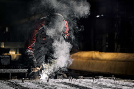 A strong man is a welder in uniform, welding mask and welders leathers, a metal product is welded with a welding machine at the construction siteの写真素材