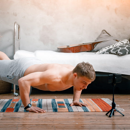 The young man goes in for sports at home. Cheerful sportsman with blond hair does push-ups, shooting...