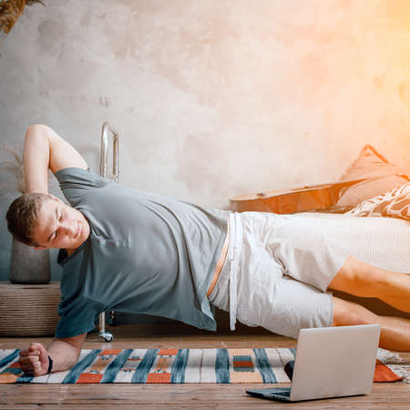 The young man goes in for sports at home. Cheerful sportsman with blond hair holds the plank on the side and and watches a movie and studies from a laptop in the bedroomの写真素材