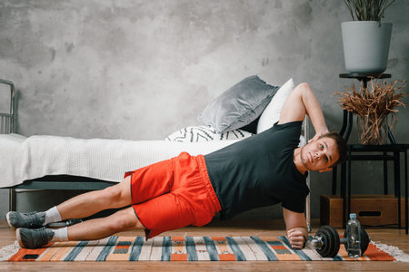 Young man goes in for sports at home, training online. The athlete keeps the plank on the side in...