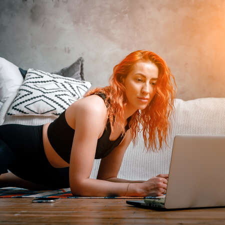 A young woman goes in for sports at home, online workout. The athlete stretching, meditating, sitting on a twine in the bedroom, in the background there is a bed, a vase, a carpet.の写真素材