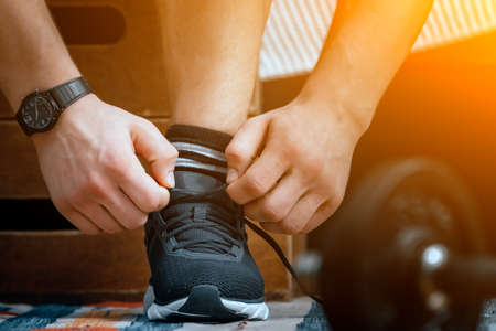 Close-up of a man tying shoelaces in black sports sneakers, a sports black watch on his hand, and metal dumbbells nearbyの写真素材