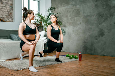 Young women go in for sports at home, workout online. Two athletes are makes lunges in the bedroom, in the background there is a bed, a vase, a carpet.の写真素材