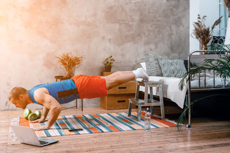 A young man goes in for sports at home, online workout. The athlete shakes biceps, do push-ups on stool in the bedroom, in the background there is a bed, a vase, a carpet.の写真素材