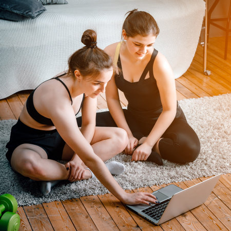 Young women go in for sports at home, workout online. Two athletes are stretching, meditating, discussing workout in the bedroom, in the background there is a bed, a vase, a carpet.の写真素材