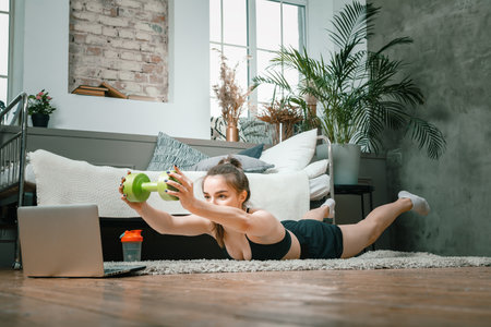 The young woman goes in for sports at home. Cheerful sportsman with black hair holds the plank with dumbbells and watching movie on laptop in the bedroomの写真素材
