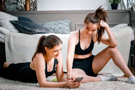 Two women friends at home chatting, smiling and playing sports. Cheerful athletic woman chatting, looking at the phone in the bedroomの写真素材
