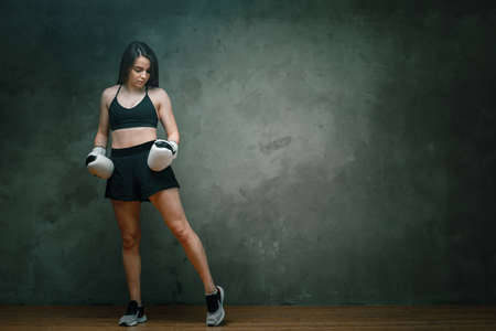 Young athletic woman boxer in shorts, short top and boxing gloves posing on dark gray background. Full length. Wall in background.の写真素材