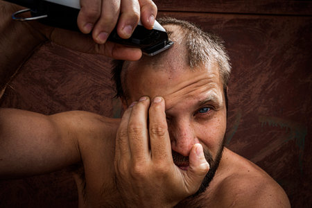 Close-up portrait of handsome shirtless man shaving his head with an electric razor against brutal...