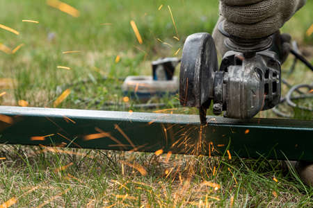 Close-up on the sides fly bright sparks from the angle grinder machine. A young male welder in a working gloves grinds a metal product with angle grinder on the grass in the villageの写真素材