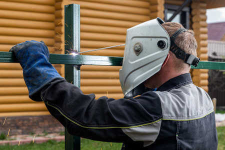 Close up of a young man welder in uniform, welding mask and welders leathers, weld metal with a...