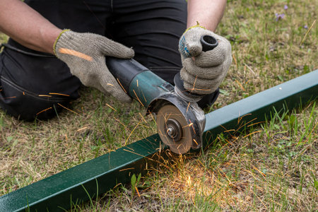 Close-up on the sides fly bright sparks from the angle grinder machine. A young male welder in a working gloves grinds a metal product with angle grinder on the grass in the villageの写真素材