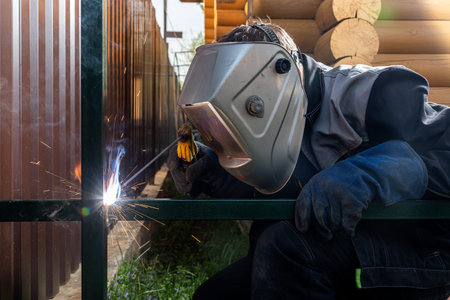 Close up of a young man welder in uniform, welding mask and welders leathers, weld metal with a...