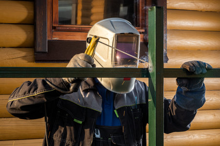 Man welder in welding mask, building uniform and protective gloves brews metal welding machine on street construction, in the background country house in summer dayの写真素材