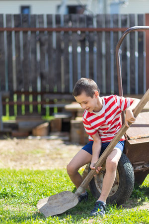 A little cheerful boy sits on a garden wheelbarrow and holds a shovel in his hand in the garden of a...